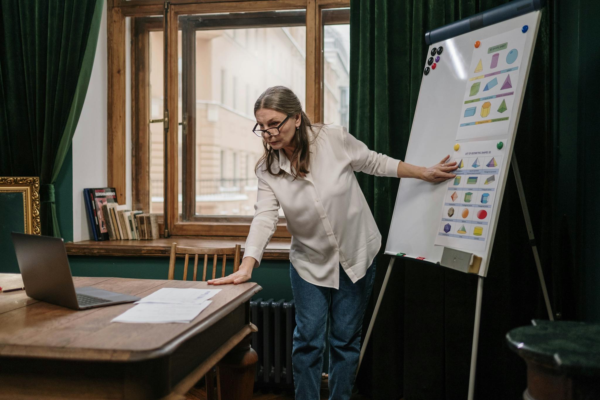Senior woman educator teaching geometry indoors with flip chart and laptop.